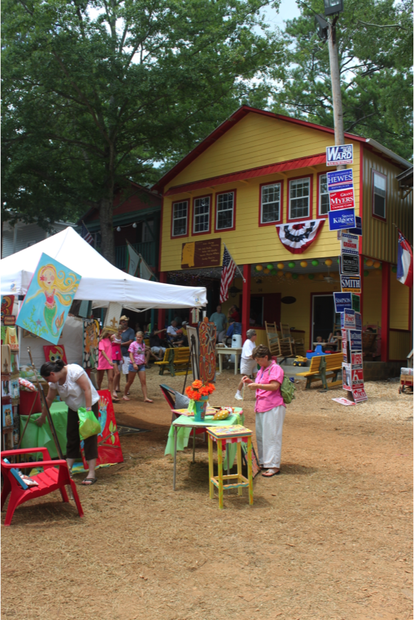 A yellow Neshoba County fair cabin stands in the back with a white tent and a lot of people walking and gathering in front of it.